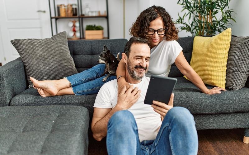 woman sits on couch with a cat in her lap while she looks over her husband's shoulder at their tablet