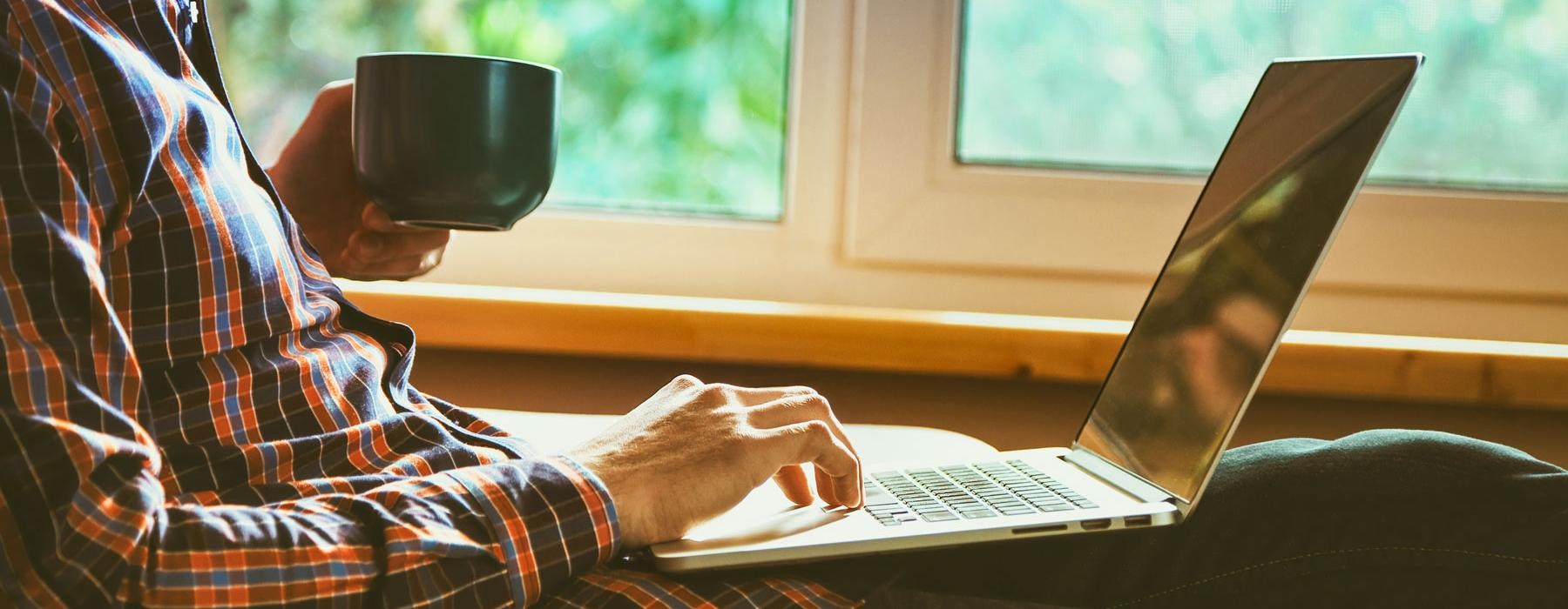 man works at a laptop while looking out the window with a cup of coffee in his hand